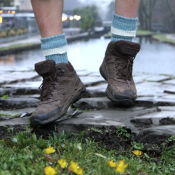A person wearing brown hiking boots and blue-and-cream ribbed wool socks is sitting on a weathered wooden fence above a muddy path beside a canal. Their legs are slightly lifted off the ground, and they are dressed in black rolled-up trousers. The background includes a stone pathway, green grass, and a calm waterway lined with trees on a misty day.